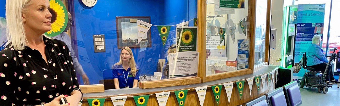The patient registration desk at Bury Care displays Sunflower bunting. Two white females with blonde hair are present. One wears a nurses uniform and sits behind the desk. 