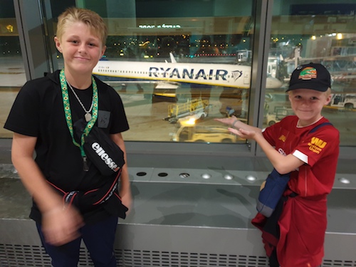 Two young white boys at the airport. They look happy. One wears a Hidden Disabilities Sunflower lanyard and there is an aeroplane on the tarmac visible through the window. 