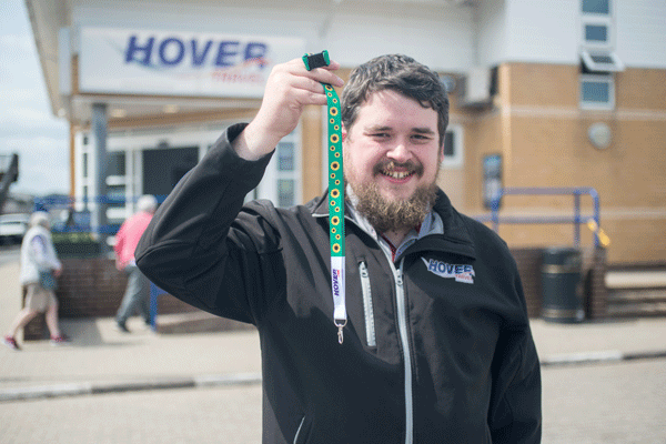 A man with dark hair and wearing a dark top is holding a green lanyard with yellow sunflower with the Hovertravel logo printed on a white section 