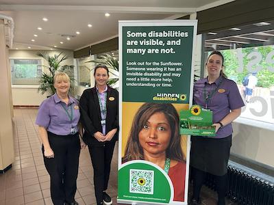 Three white, female employees of Centre Parcs flank a Hidden Disabilities Sunflower pull up banner. One of the people hold a green box with the Sunflower logo on containing lanyards. 