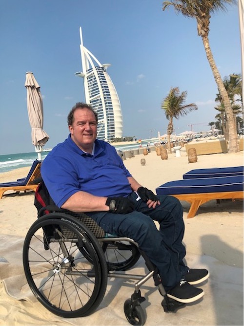 Fred Maahs in his wheelchair on a beach in Dubai. The Burj Al Arab is in the background. 