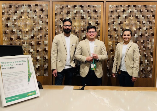 The hotel reception at Sudima Hotel. Three employees stand behind a desk. The desk has a Hidden Disabilities poster in a frame on it.  