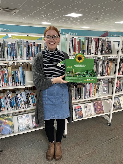 A young white woman wearing glasses stands in a library holding a box of hidden disabilities sunflower lanyards 