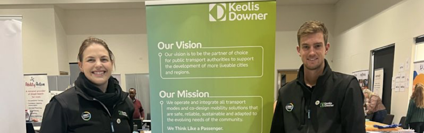 Two people from Keolis Downer stand either side of a \Keolis Downer promotional banner in a conference hall
