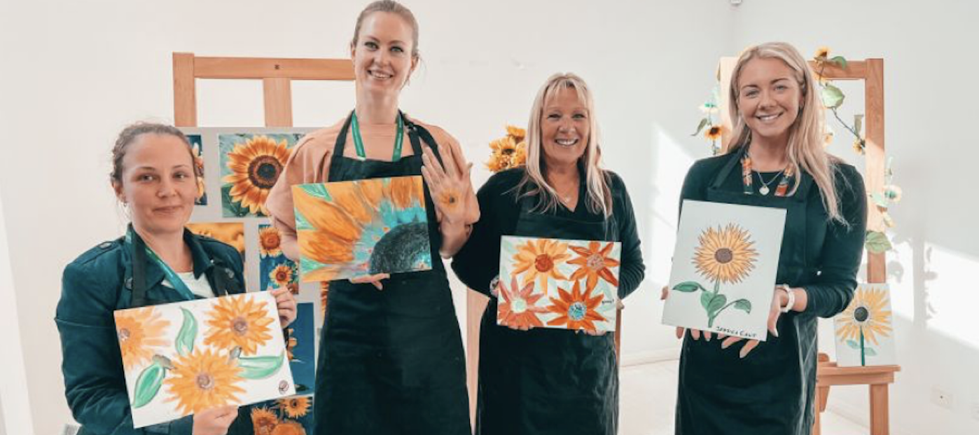 A group of white women have painted some sunflowers and are showing their art to the camera