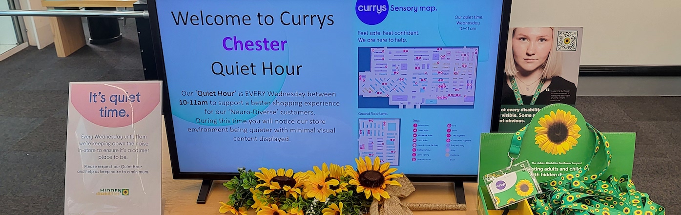 A table top with a digital screen that reads: Welcome to Currys Chester quiet hour. There are Sunflowers, a box of lanyards and information leaflets also on the table