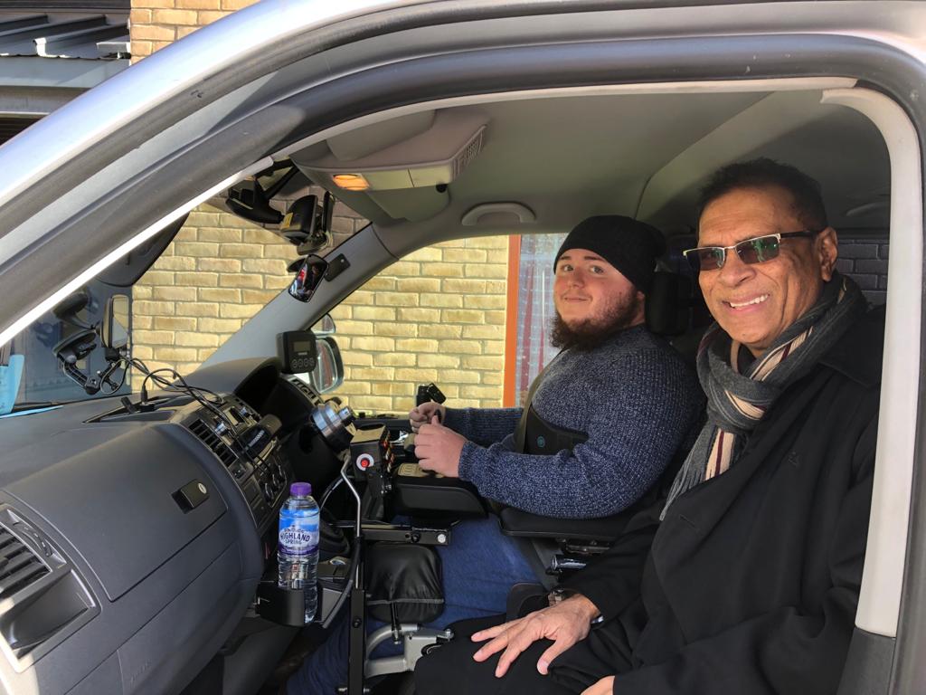 A specially adapted vehicle with a yound man with disabilities in the driver's seat. Driving instructor Eshan sits next to him, smiling at the camera. 