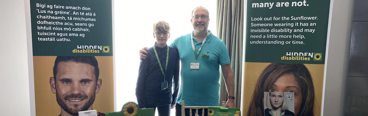 Tristan stands at the Sunflower table top booth with his son. There are Hidden Disabilities banners each side of them and they stand in front of a window