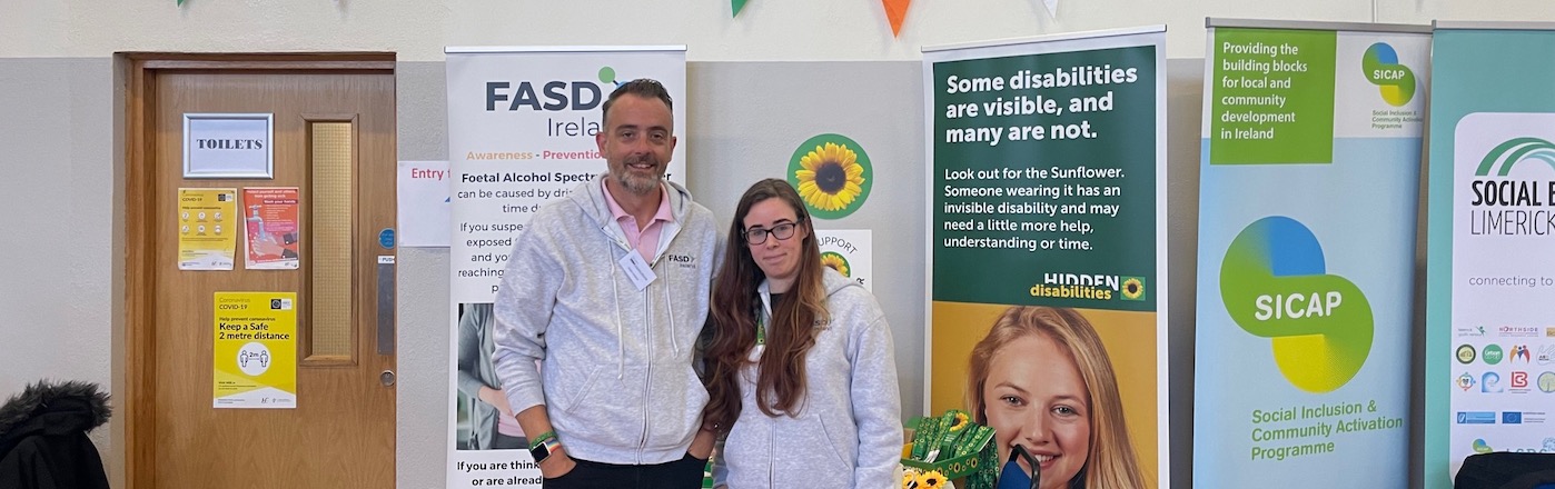 Scott and Maggie, a white man and a white woman, stand in front of a hidden disabilities Sunflower and FASD display