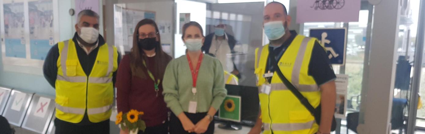 Four people stand in an airport terminal. They all wear surgical masks. the two men wear high vis jackets. One of the women wears a Sunflower lanyard