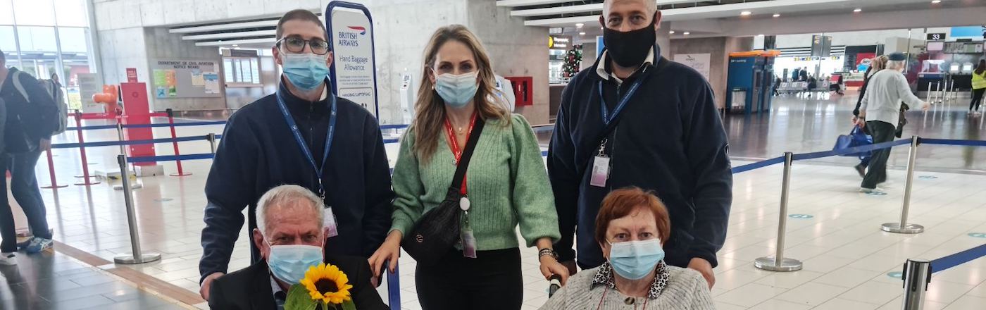 the inside of an airport terminal. Two men flank a woman, they all wear surgical masks. There are an elderly man and woman in front of them who are in wheelchairs. The elderly man carries a Sunflower 