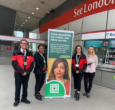 Four employees at the London Cable car stand next to a Hidden Disabilities Sunflower pull up banner. They are standing in front of the cable car ticket office in the cable car terminal