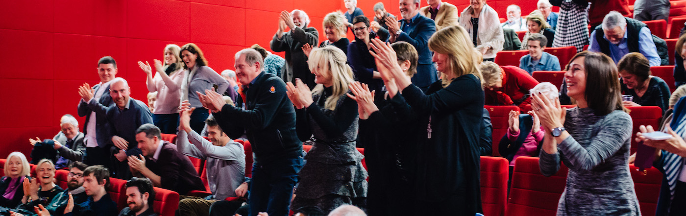 A red theatre. Audience members clap. Some are standing. 