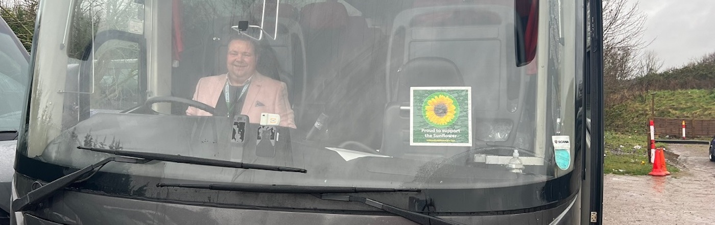 Darrly sits behind the wheel of a black coach. There is a Sunflower sticker on the windscreen