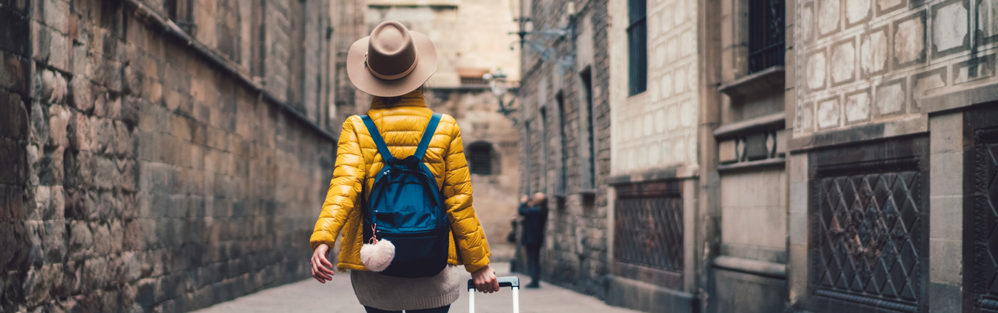 A woman in a yellow puffer jacket walks down an old street with a blue case behind her. She has her back to us and wears a brown sun hat