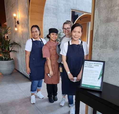  Four female members of Sudima staff stand in the hotel lobby. There is a Hidden Disabilities Sunflower poster on display.  