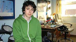 A picture of Greg Storey, a young white male. He wears a green jumper and sits in his bedroom. A drum kit and desk are visible in the background. 