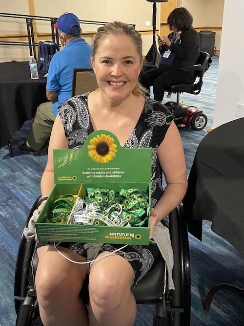A young, blonde, white woman sits in a wheel chair. She holds a box of Sunflower lanyards 