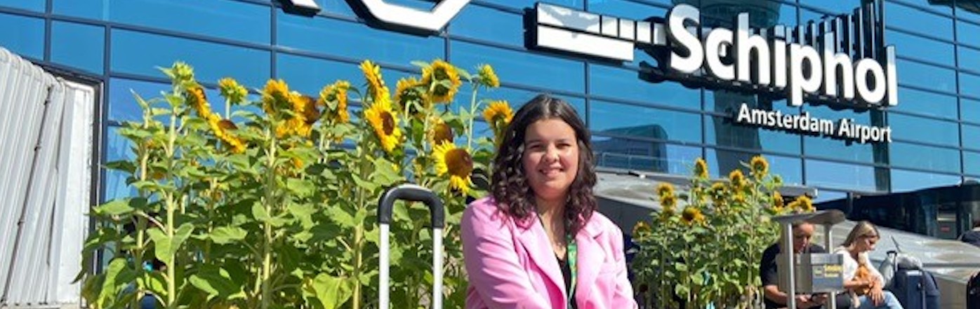 A female Sunflower wearer with long dark hair, wearing a pink jacket, sits outside of Schiphol airport. There are Sunflowers growing around her. 